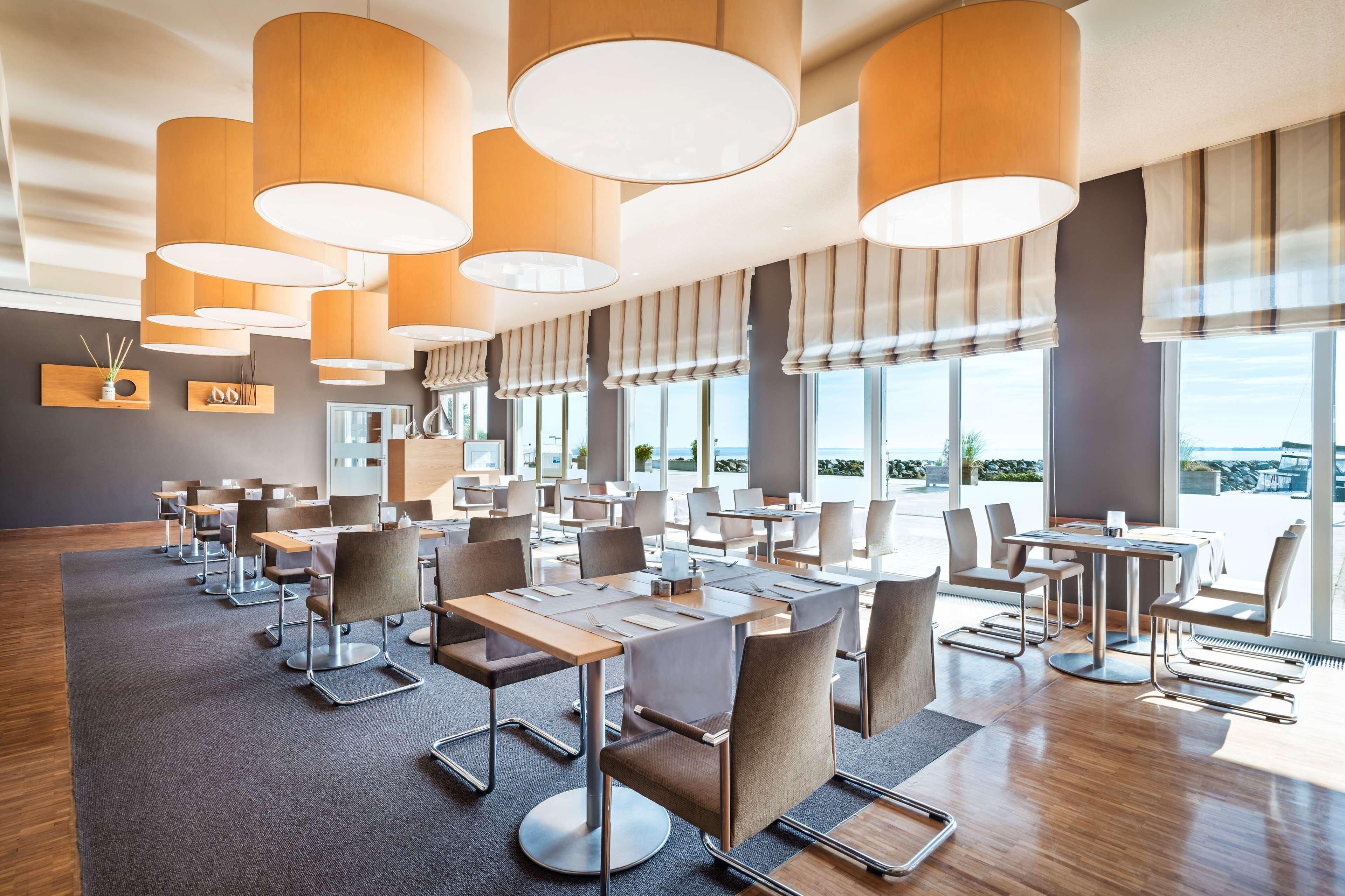 A modern dining area with multiple tables and chairs, large windows, and hanging pendant lights.