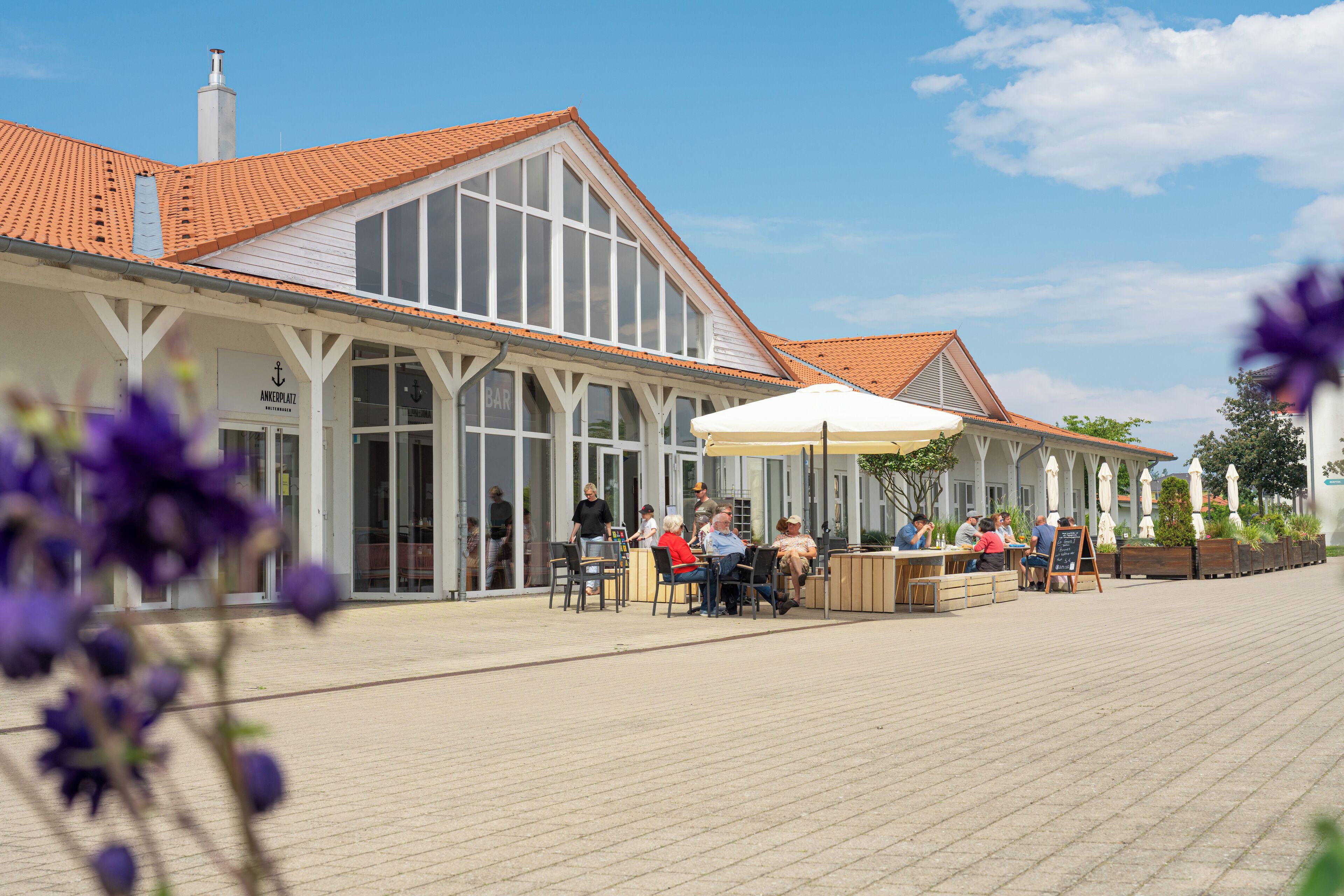 A building with a tiled roof and large windows, featuring outdoor seating with umbrellas and a sign that reads "Anchor Plaza Bar".