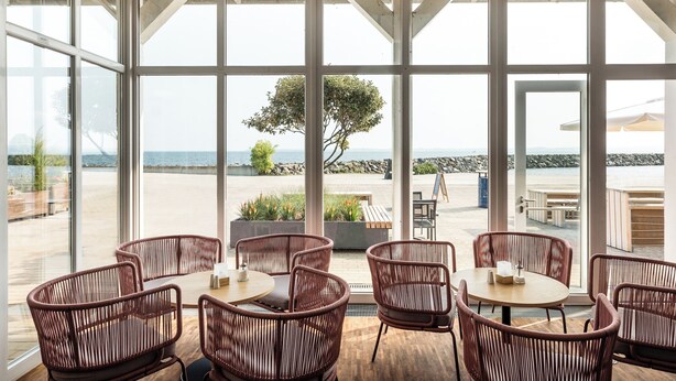 A spacious outdoor seating area with wicker chairs and tables, overlooking a rocky shoreline and the sea.