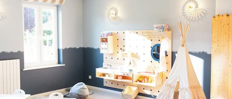 Ein heller, gut beleuchteter Raum mit Fenster, einem Spielbereich mit einem weißen Zelt und einem hölzernen Spielhäuschen.