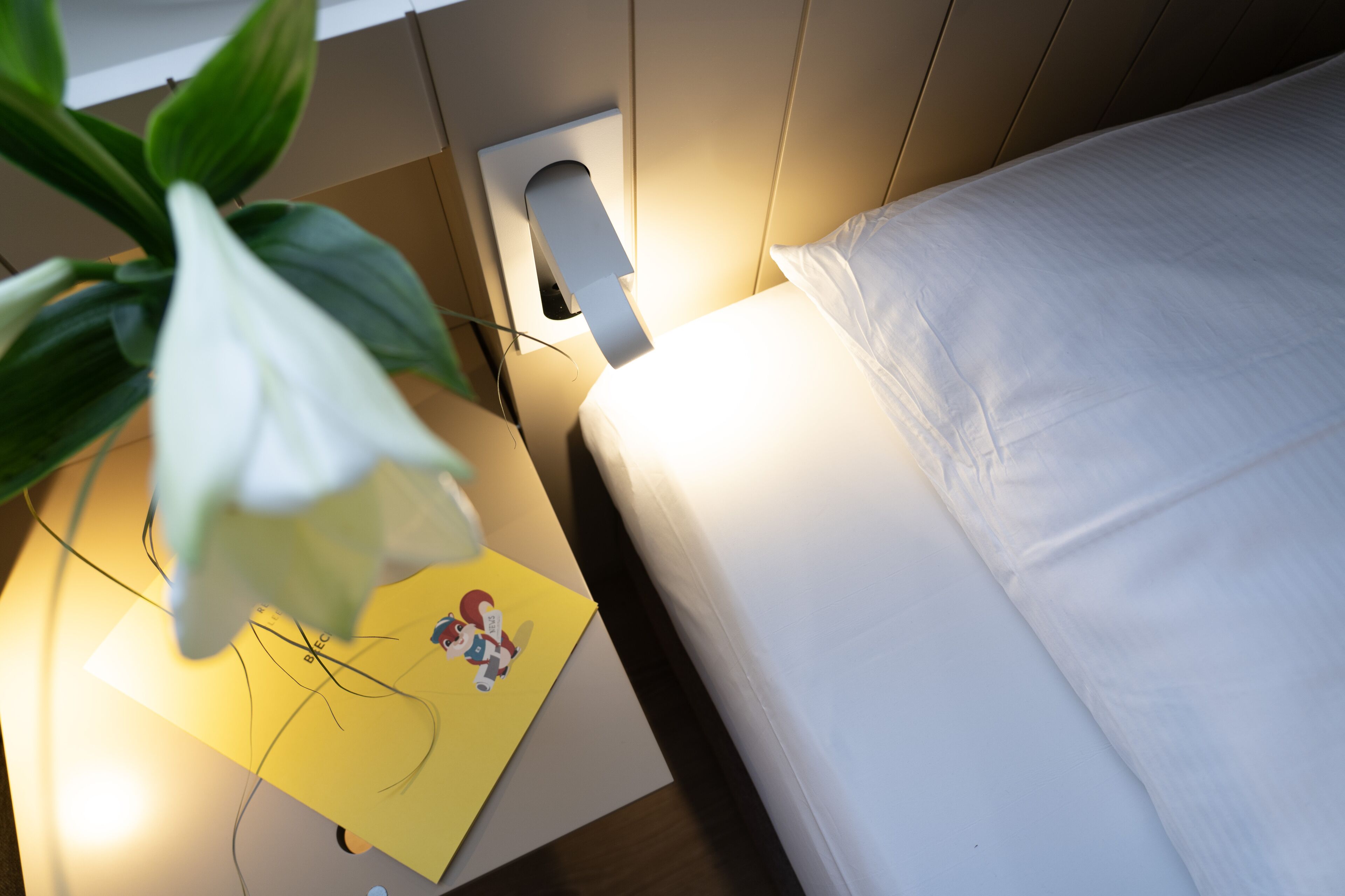 A neatly made bed with white bedding, a bedside lamp, and a yellow note with a cartoon character.