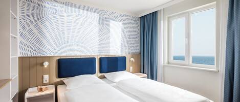 Un lit double avec du linge de lit blanc, une table de chevet en bois, une lampe fixée au mur et une fenêtre donnant sur la mer.