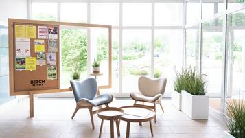 A modern lobby with a corkboard displaying various notices, two armchairs, a small table, and potted plants.