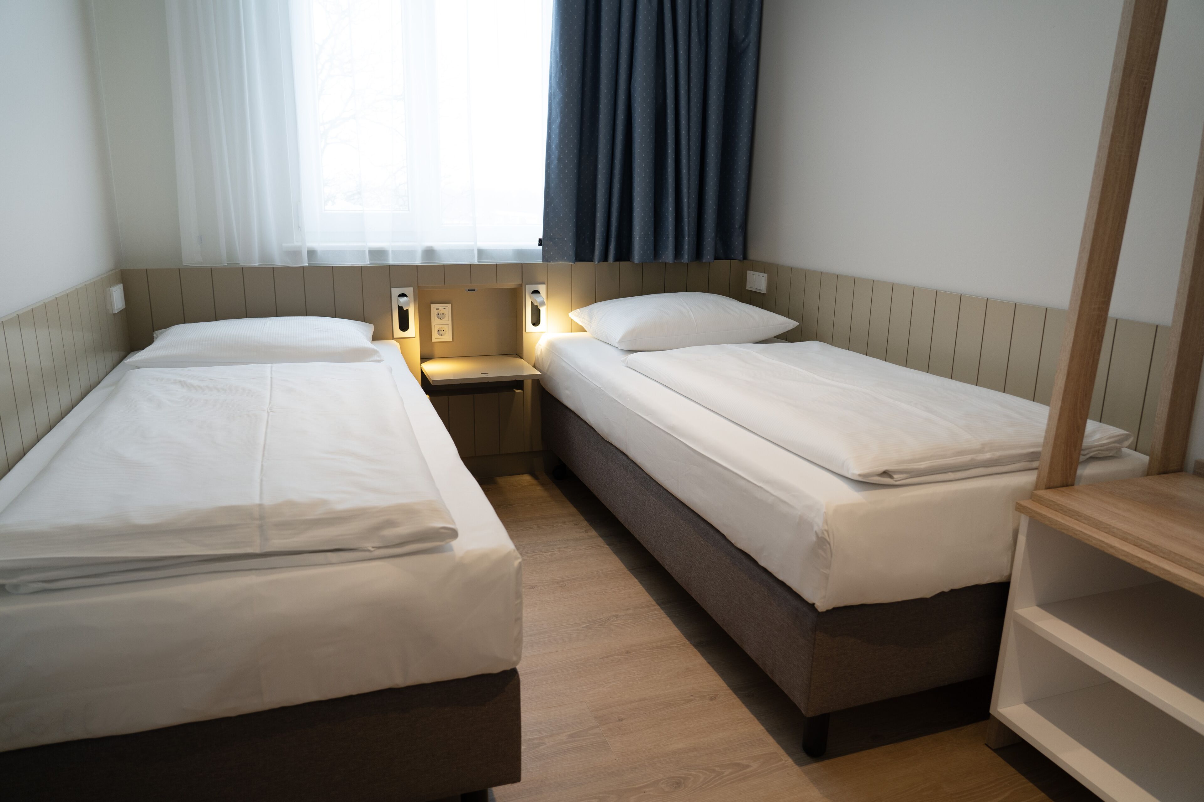 Quarto de hotel com duas camas de solteiro, uma pequena mesa de cabeceira com abajur, um criado-mudo de madeira e uma janela com cortinas.