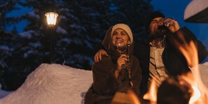 A couple sitting by a campfire with a snow-covered landscape and a street lamp in the background.
