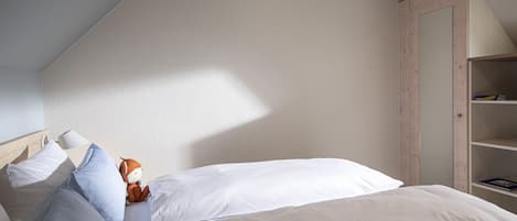 Un dormitorio con techo inclinado, una cama con ropa de cama blanca, una mesita de noche de madera y una lámpara, además de un peluche.