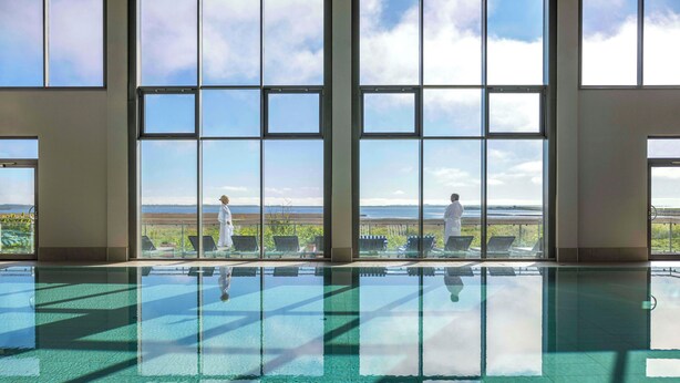 A spacious indoor swimming pool with clear glass walls, offering expansive views of the surrounding landscape. - TUI BLUE Sylt (Sylt)