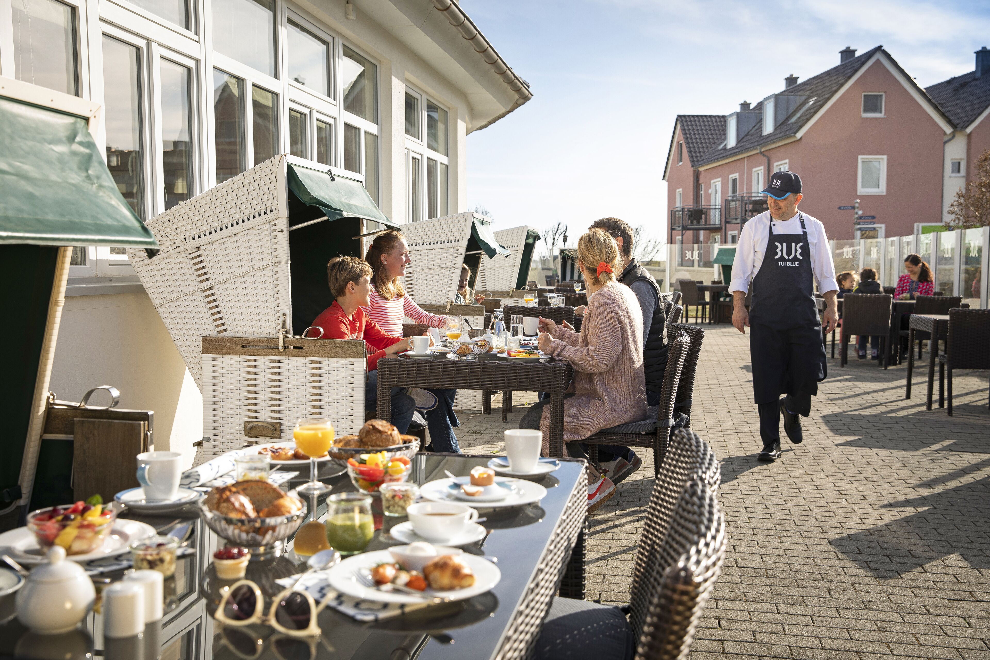 An outdoor dining area with wicker furniture, a table set with breakfast items, and a person walking by.