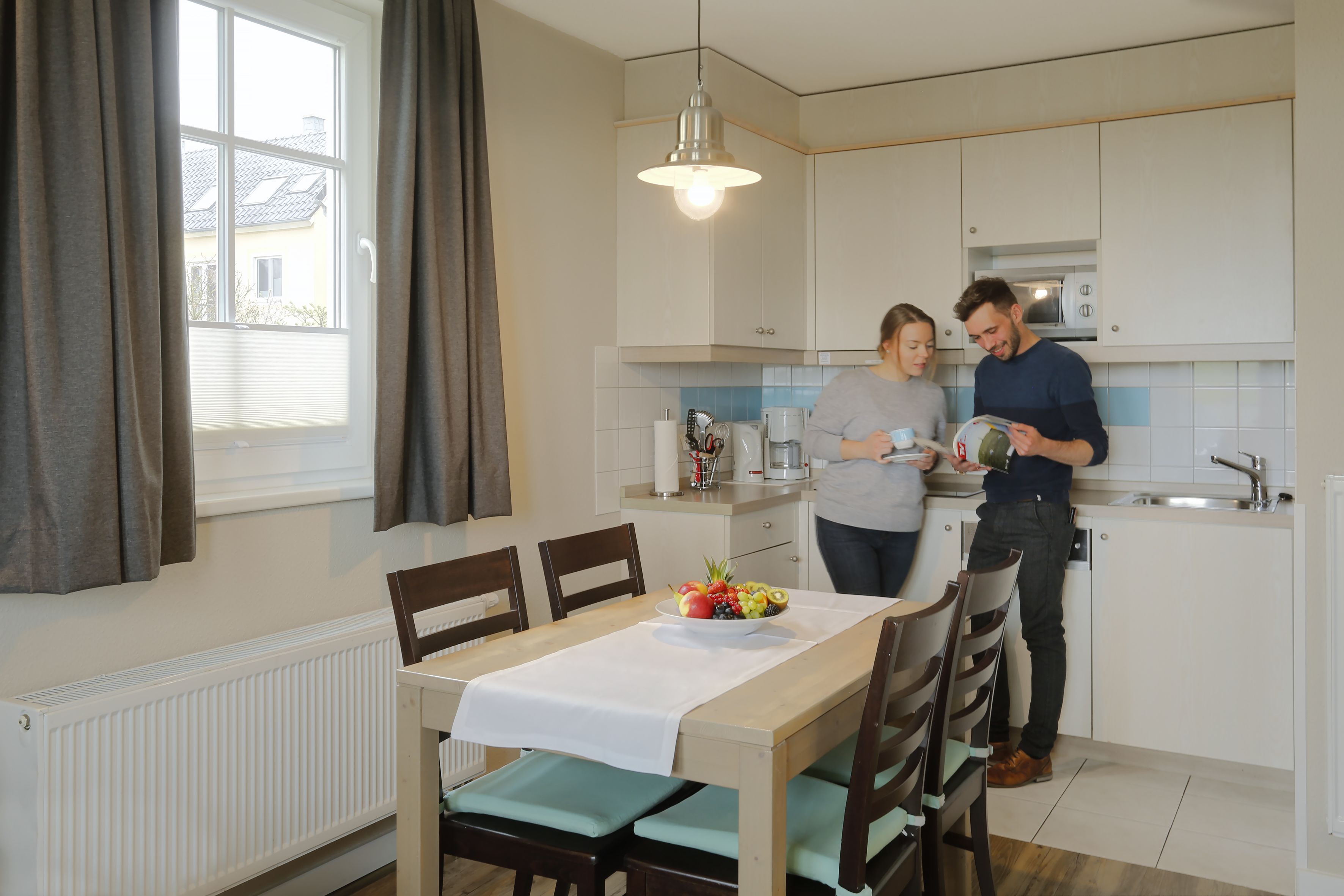 Een moderne keuken met witte kastjes, een eettafel met een fruitschaal, en een raam met gordijnen.