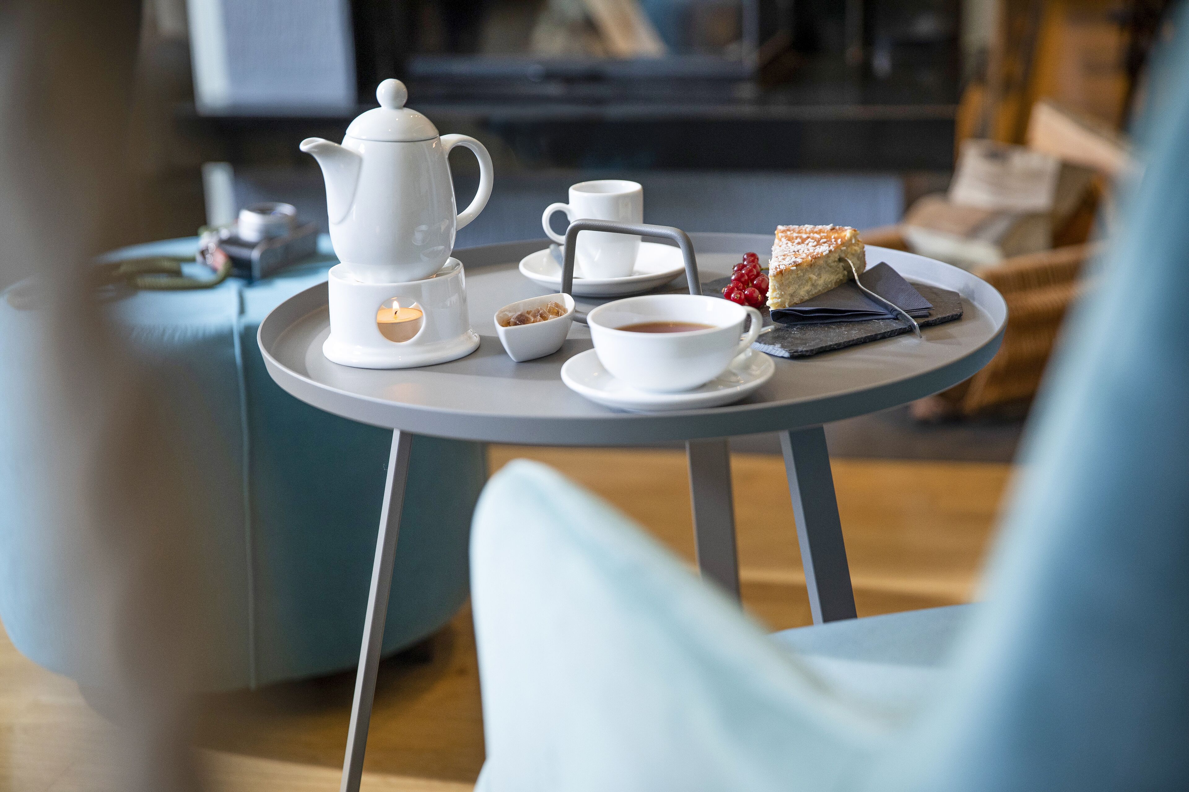 Une table ronde sur laquelle sont disposés une théière blanche, des tasses, un petit bol et une part de gâteau, le tout dans une pièce moderne avec une chaise bleue et un téléviseur en arrière-plan.