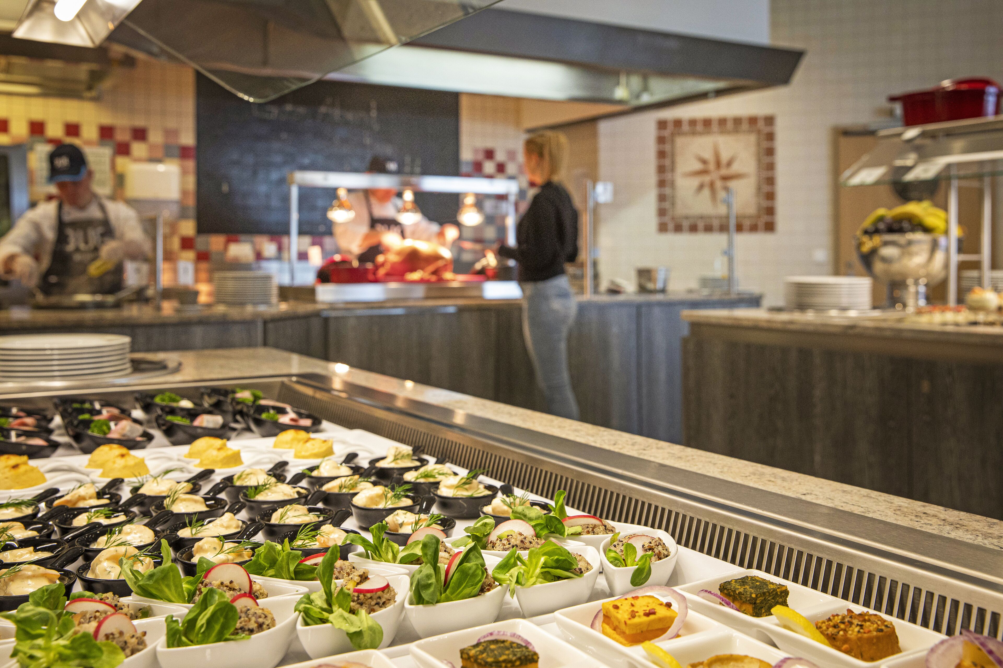 A buffet with various dishes arranged in trays, a kitchen area with chefs preparing food, and a counter with plates and utensils.
