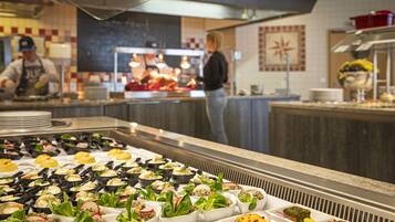 A buffet with various dishes arranged in trays, a kitchen area with chefs preparing food, and a counter with plates and utensils.