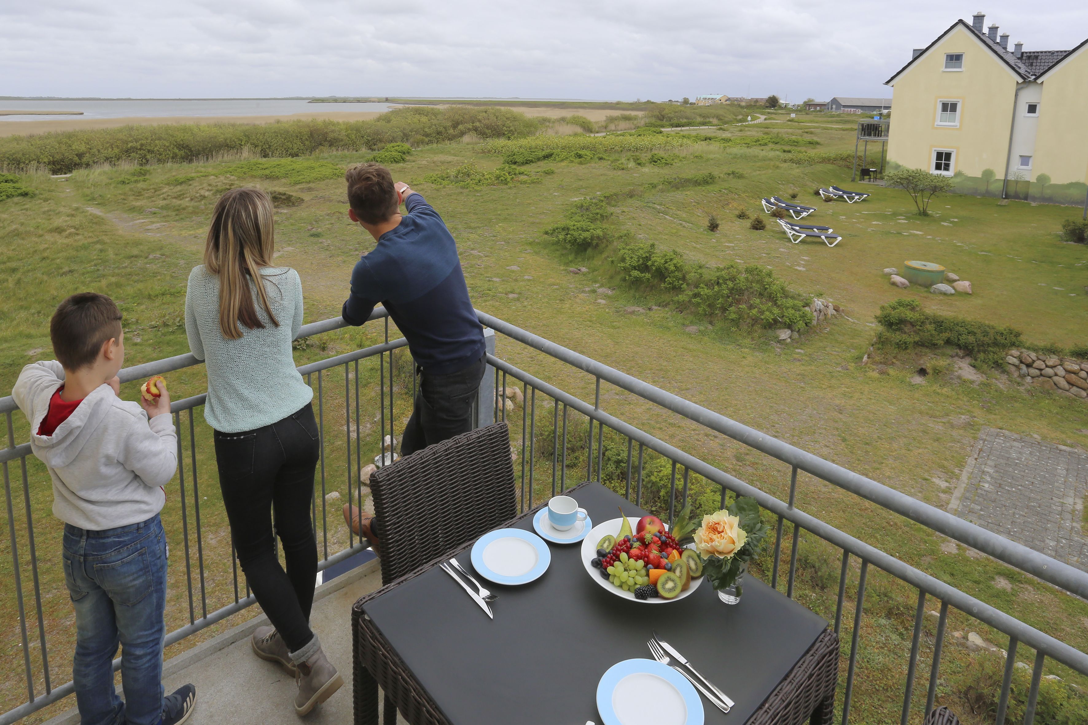 Un balcón con una mesa dispuesta para una comida, con vista a una zona verde y un edificio al fondo.