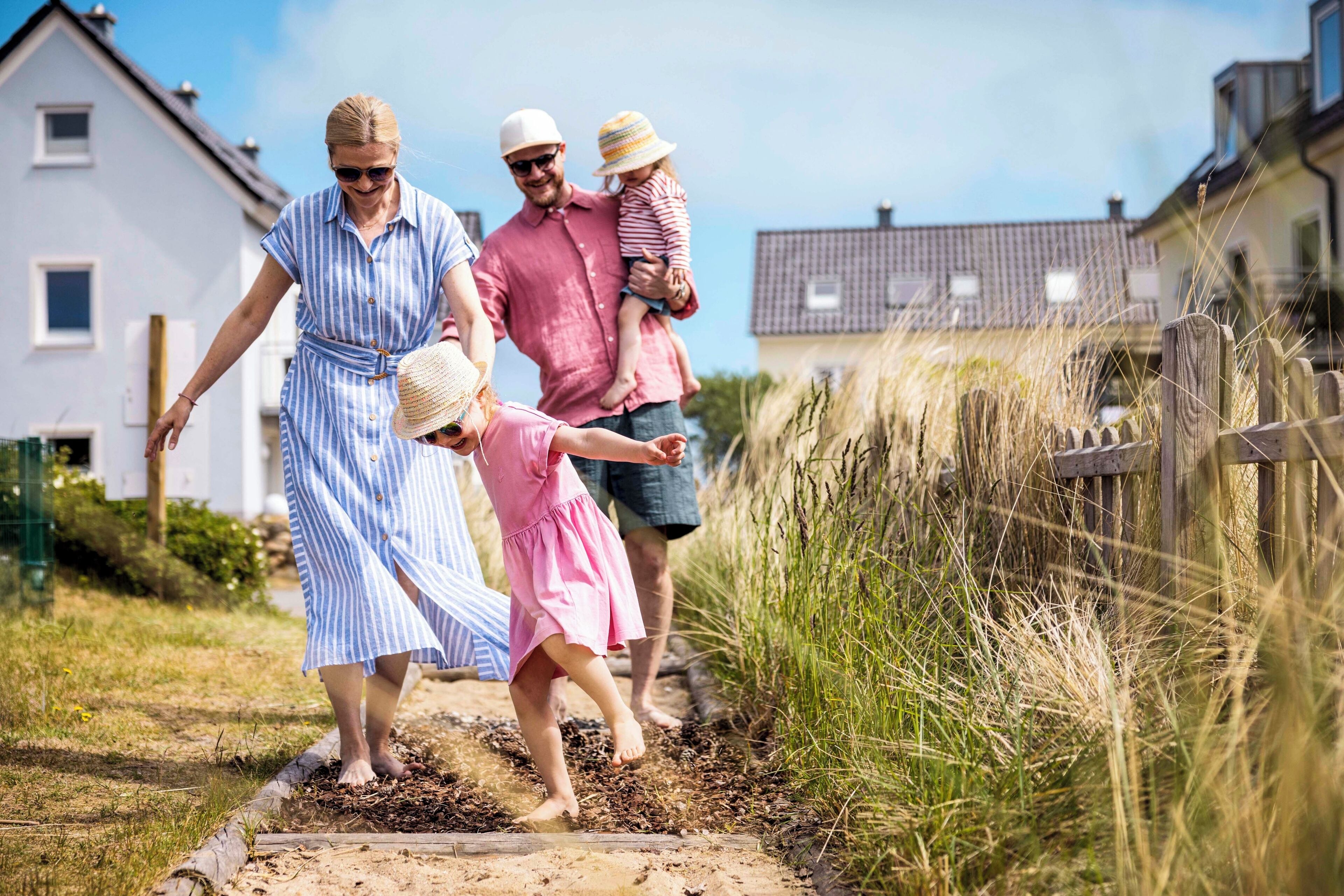 A family walking on a dirt path beside a house with a white exterior and a wooden fence.
