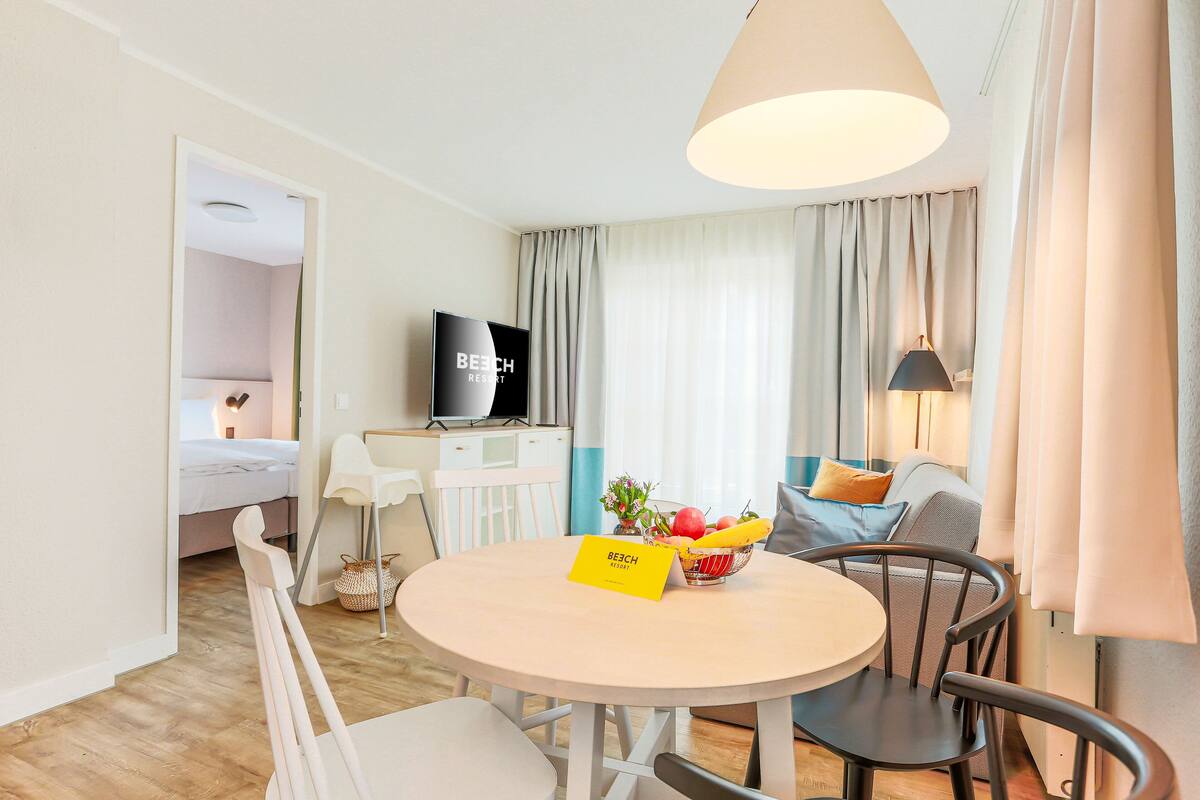 A hotel room with a bed, a TV, a dining table with chairs, a lamp, and a sign that reads "BEACH RESORT".