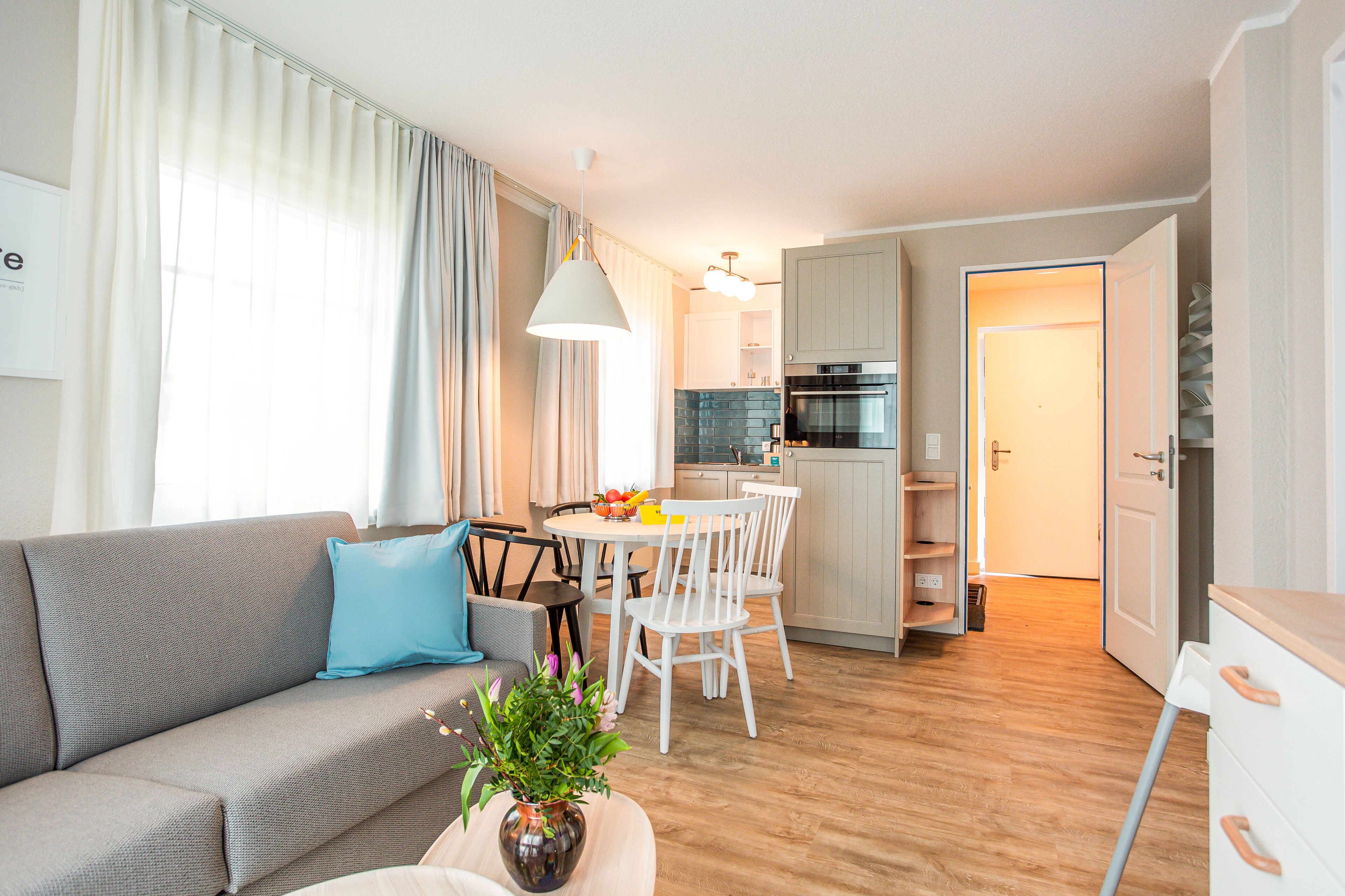 Ein modernes Wohnzimmer mit grauem Sofa, einem Esstisch mit Stühlen, einer Küchenzeile mit Herd und Schränken sowie einer Treppe, die zu einer weiteren Ebene führt.