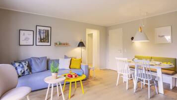 Uma sala de estar moderna com sofá, mesa de jantar com cadeiras, pequena mesa lateral com frutas e luminária.
