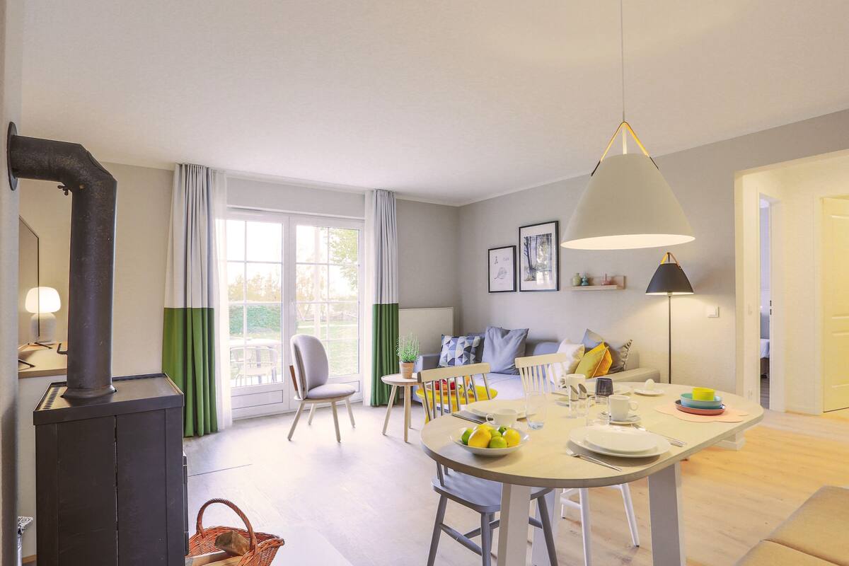 A modern living room with a sofa, a dining table with chairs, a large window with curtains, and a black stove.