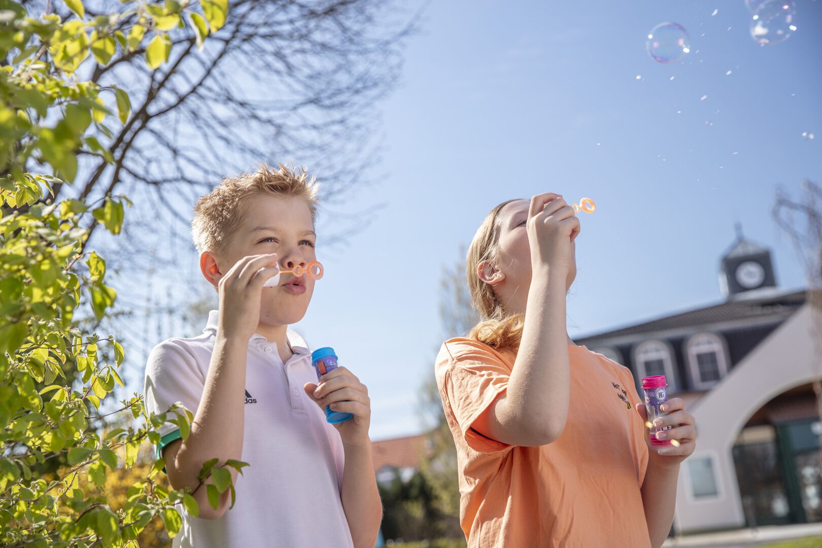 Two people blowing bubbles outdoors with a building in the background.