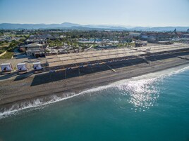 Vista aérea de un complejo turístico en la playa, con una extensa franja de arena, sillas de descanso y algunas cabañas.