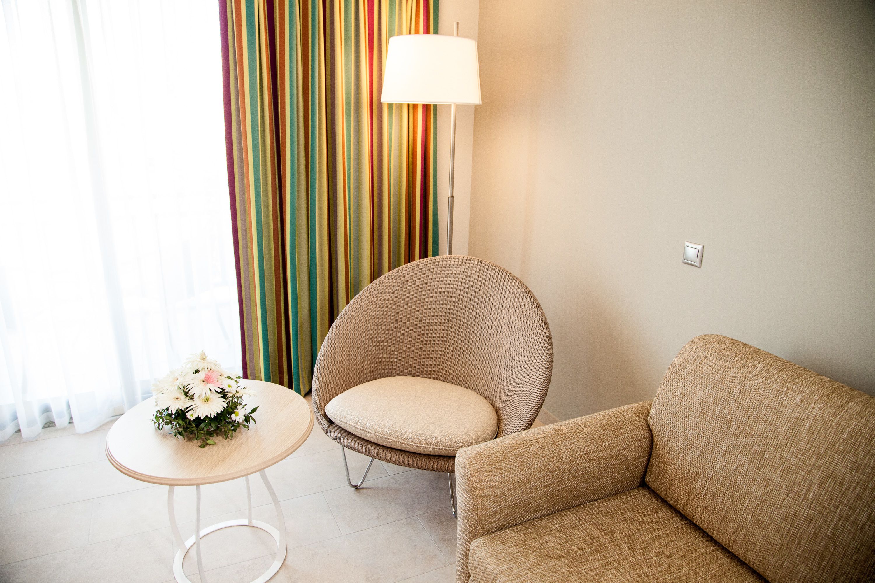 Una habitación con un sofá color beige, un sillón redondo, una mesita con un arreglo floral y una cortina a rayas coloridas.