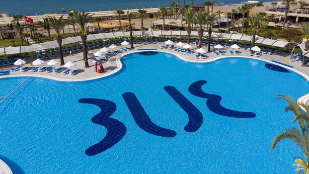 A large pool with a unique design, surrounded by lounge chairs and umbrellas, with a view of the beach and ocean. - TUI BLUE Palm Garden - All inclusive (Manavgat)