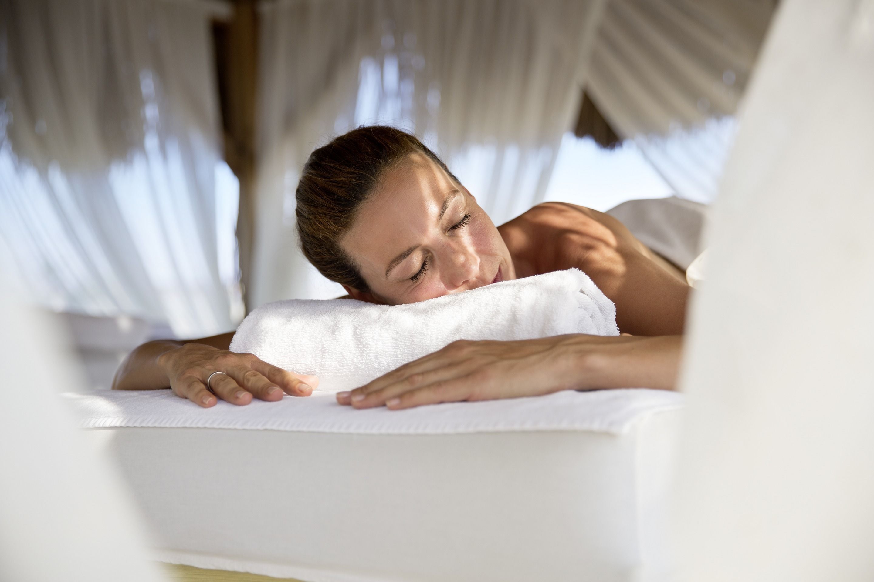 Une personne allongée sur un lit, avec un oreiller blanc et une couverture blanche, dans une pièce où sont accrochés des rideaux légers.