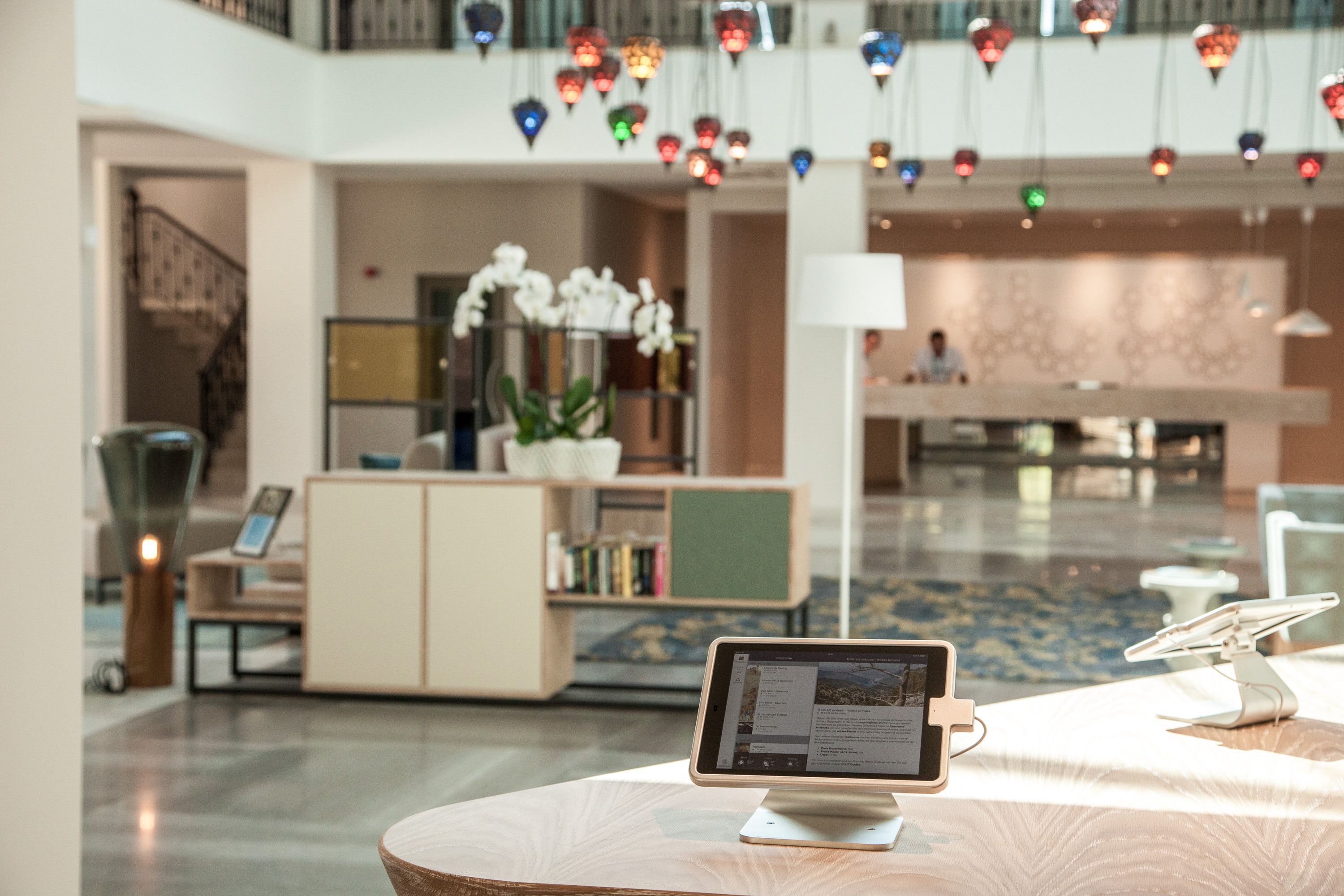 A modern hotel lobby with a reception desk, a digital kiosk, and a decorative hanging light fixture.