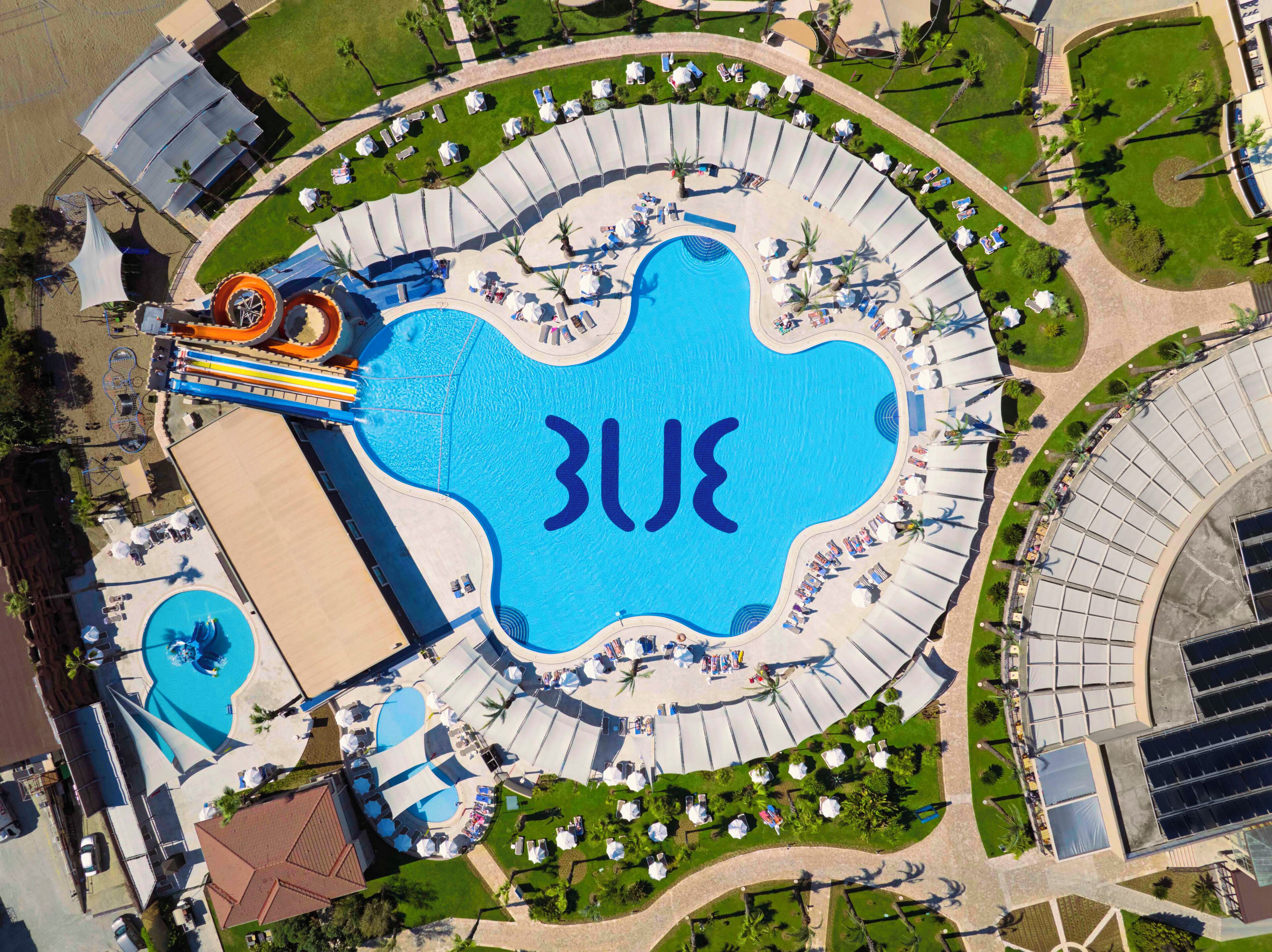 An aerial view of a resort with a large central pool, multiple smaller pools, and a water slide.