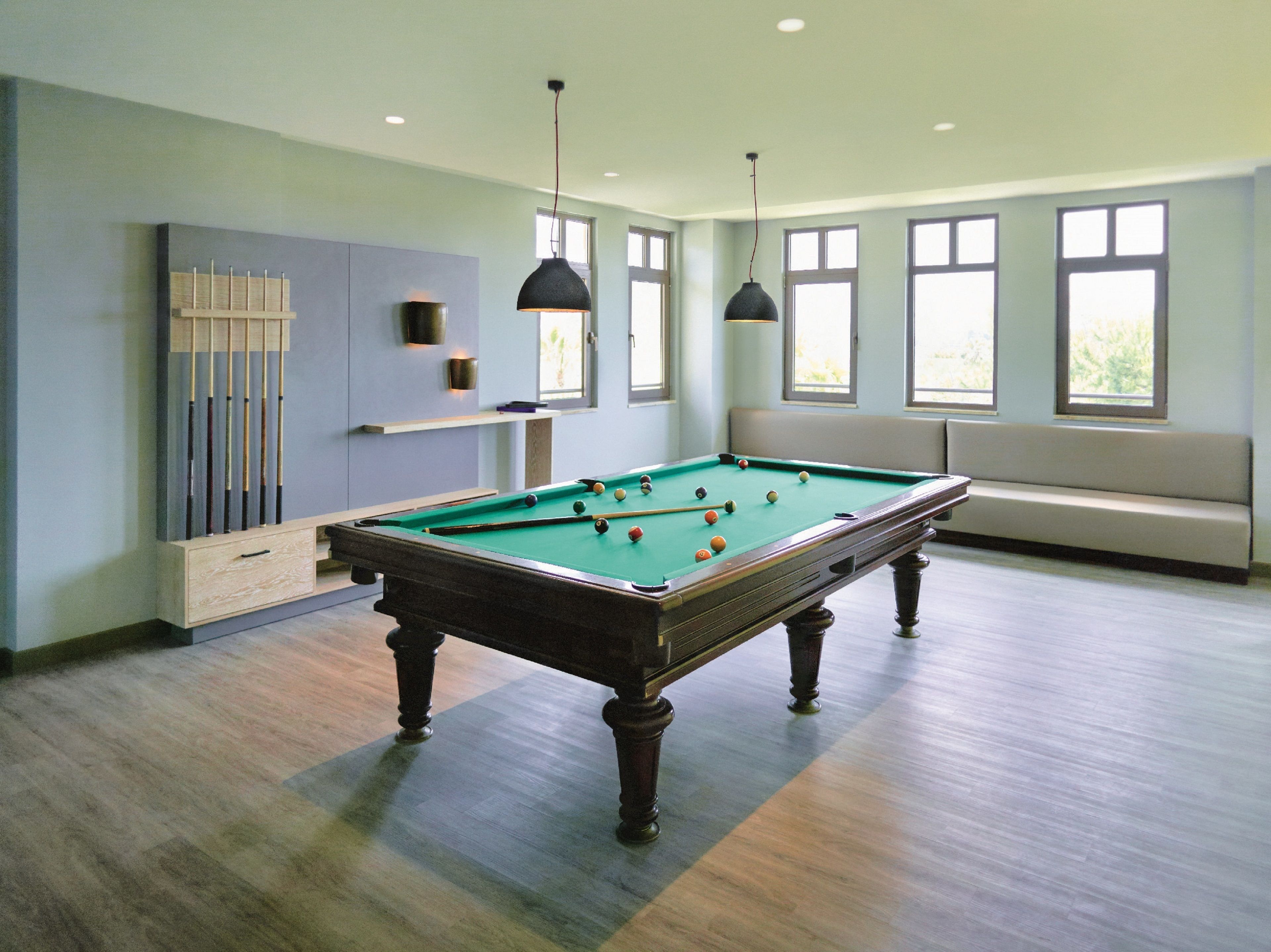 Una mesa de billar en una habitación con pared azul claro, un banco y ventanas grandes.