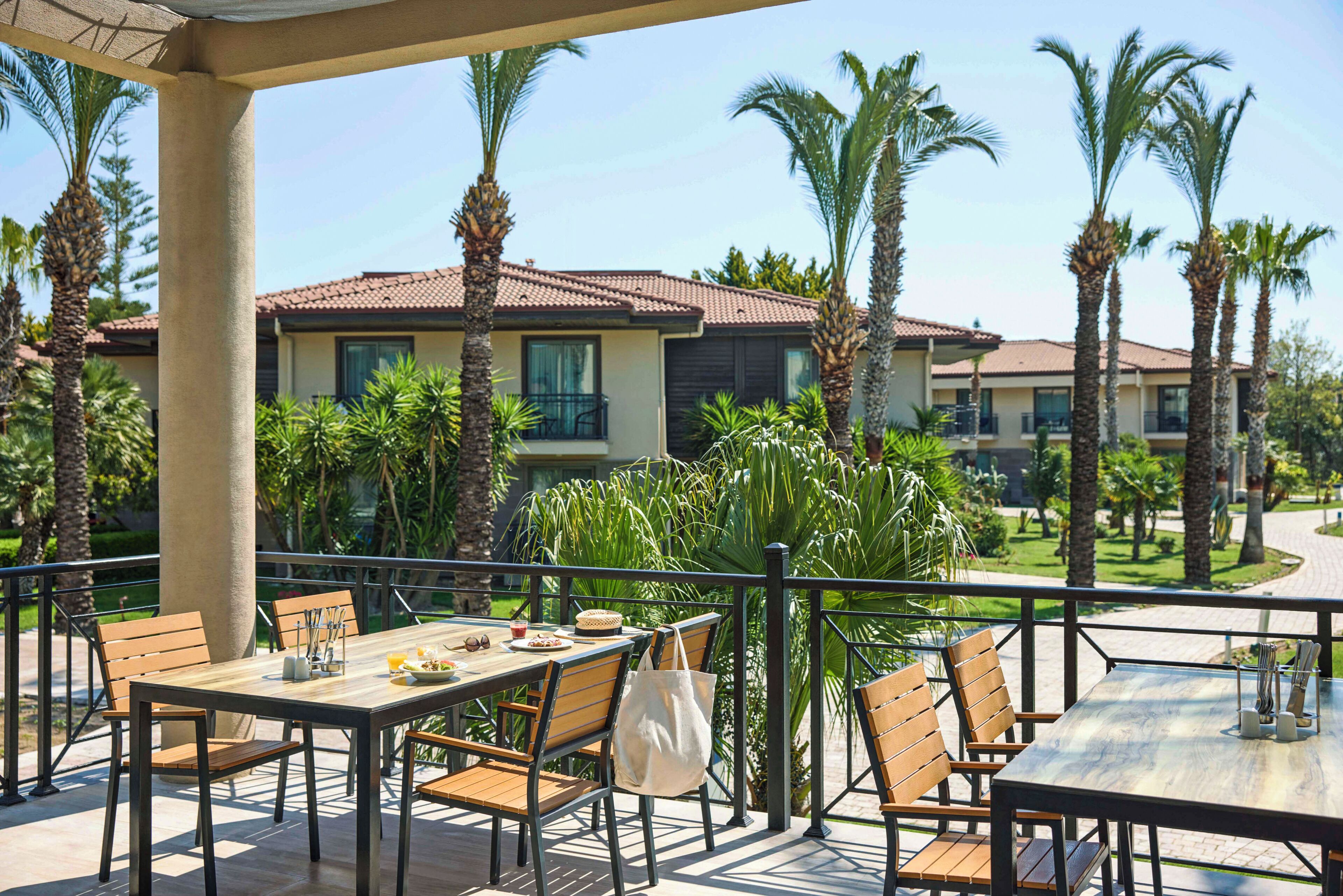 An outdoor patio with a table and chairs, surrounded by palm trees and a building with a tiled roof.