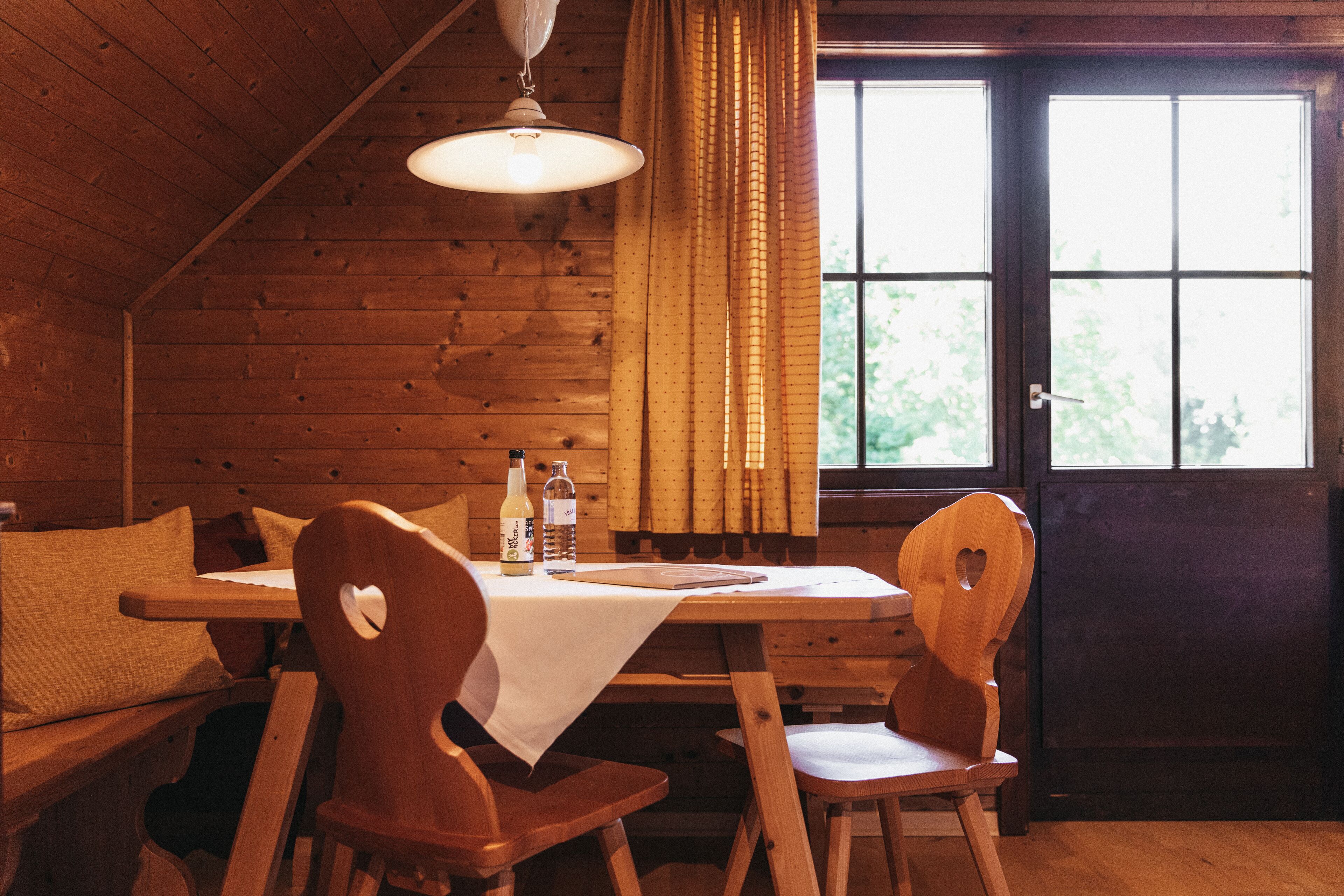A wooden cabin interior with a dining table, two heart-shaped chairs, a bottle, and a notebook.