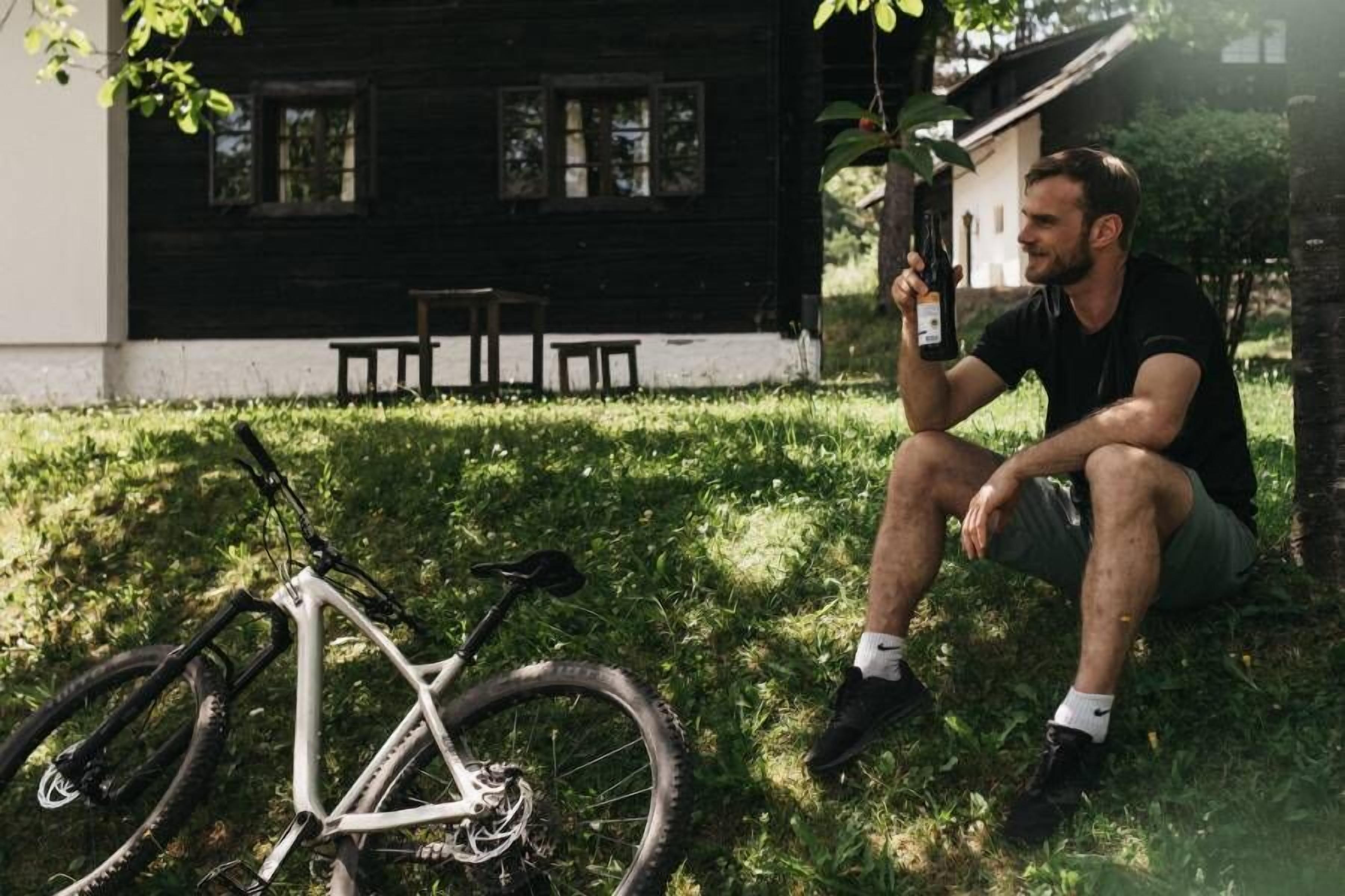 Una persona sentada sobre la hierba junto a una bicicleta de montaña blanca, con un edificio negro al fondo.