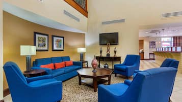 A hotel lobby with blue sofas, a coffee table, and a TV. There are framed pictures on the wall and a staircase in the background.