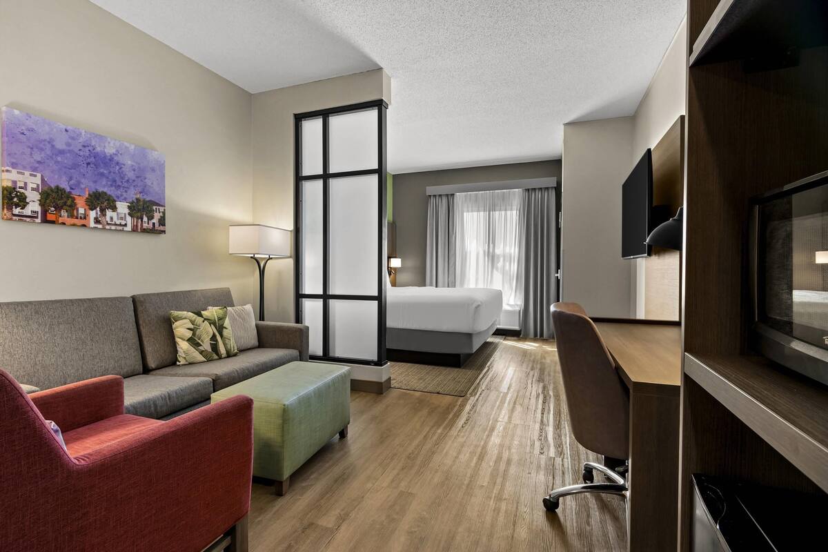 Une chambre d’hôtel équipée d’un canapé, d’un fauteuil, d’un pouf, d’un lit, d’un bureau, d’une télévision et d’un tableau au mur.
