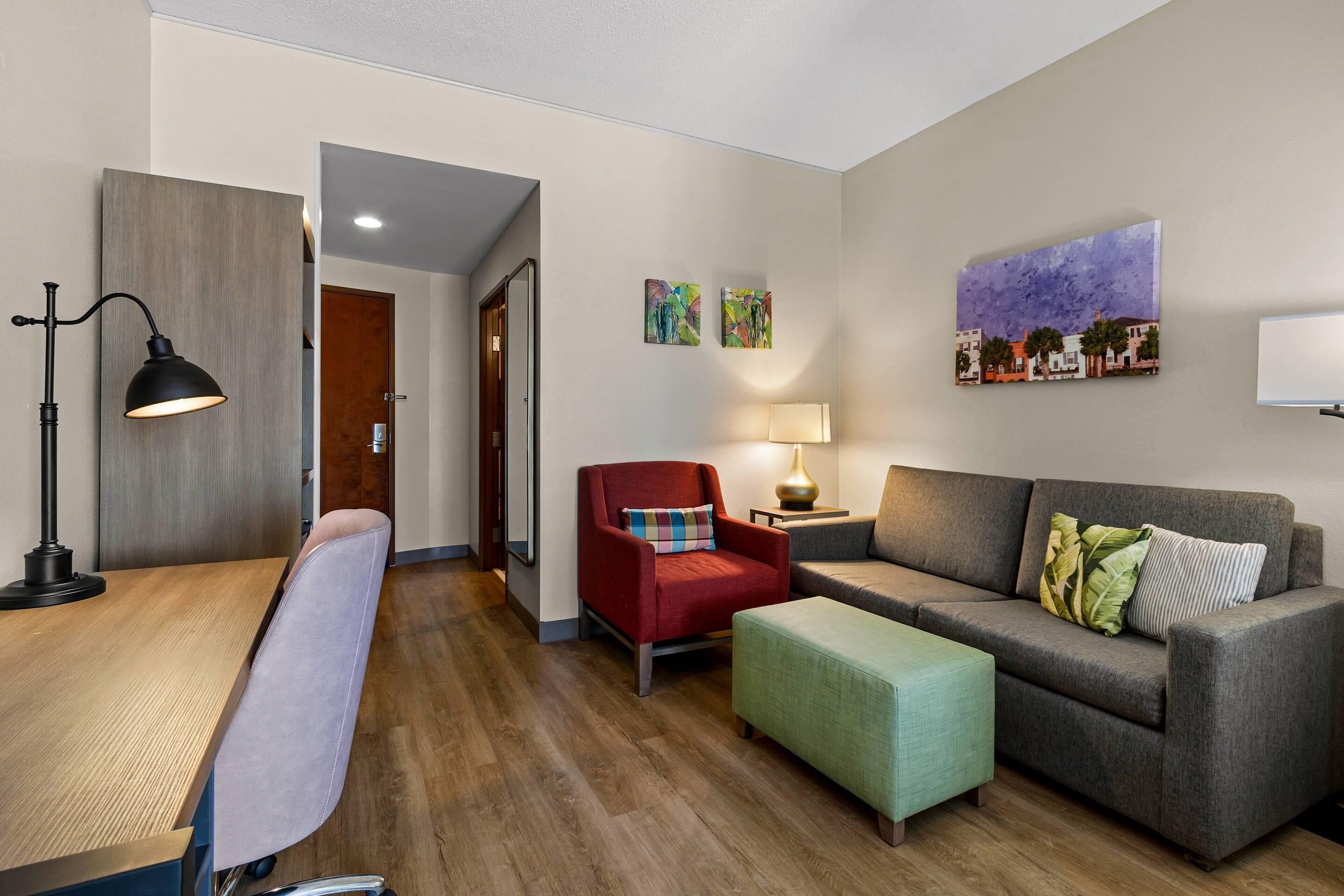 Una sala de estar moderna con un escritorio de madera, un sillón rojo, un sofá gris y un otoman verde. Hay dos lámparas de pared y un cuadro de casas.