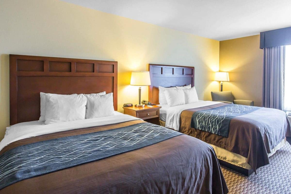 A hotel room with two beds, wooden headboards, bedside tables with lamps, and a window with curtains.