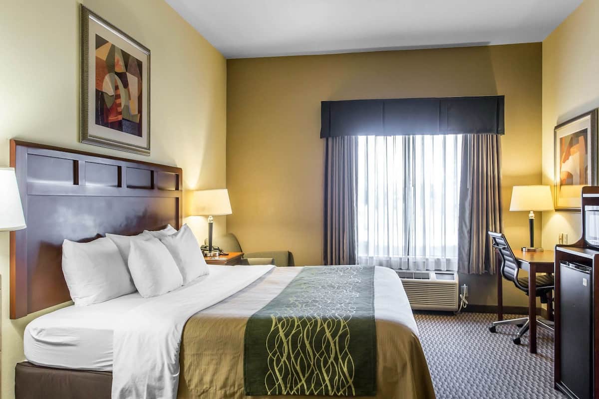 A hotel room with a bed, two bedside tables with lamps, a desk with a chair, a large window with curtains, and artwork on the walls.