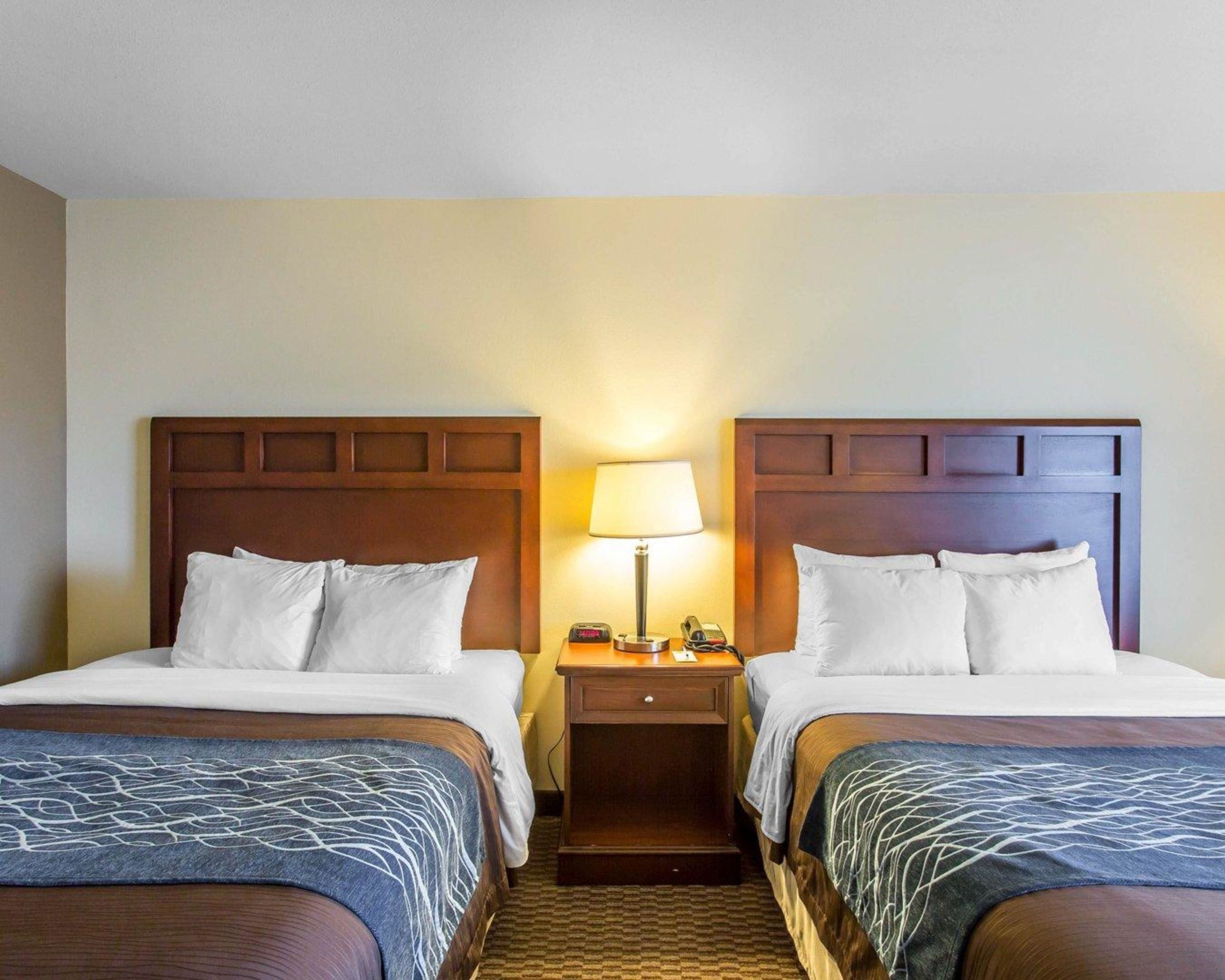 A hotel room with two beds, each with a wooden headboard, a bedside table with a lamp, and a patterned bedspread.