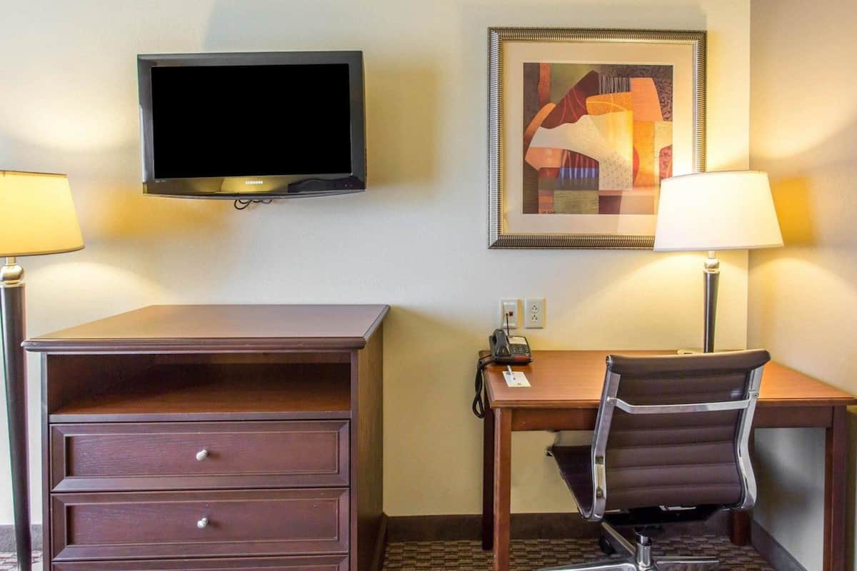 A hotel room with a flat-screen TV, a desk with a chair, a lamp, a telephone, and a framed abstract painting on the wall.