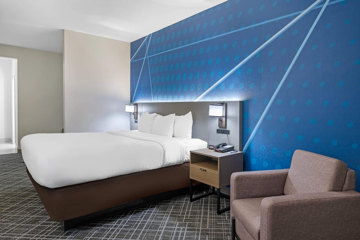 Ein Hotelzimmer mit einem großen Bett, einem Nachttisch mit Lampe, einem Sessel und einer Wand mit geometrischem Muster.