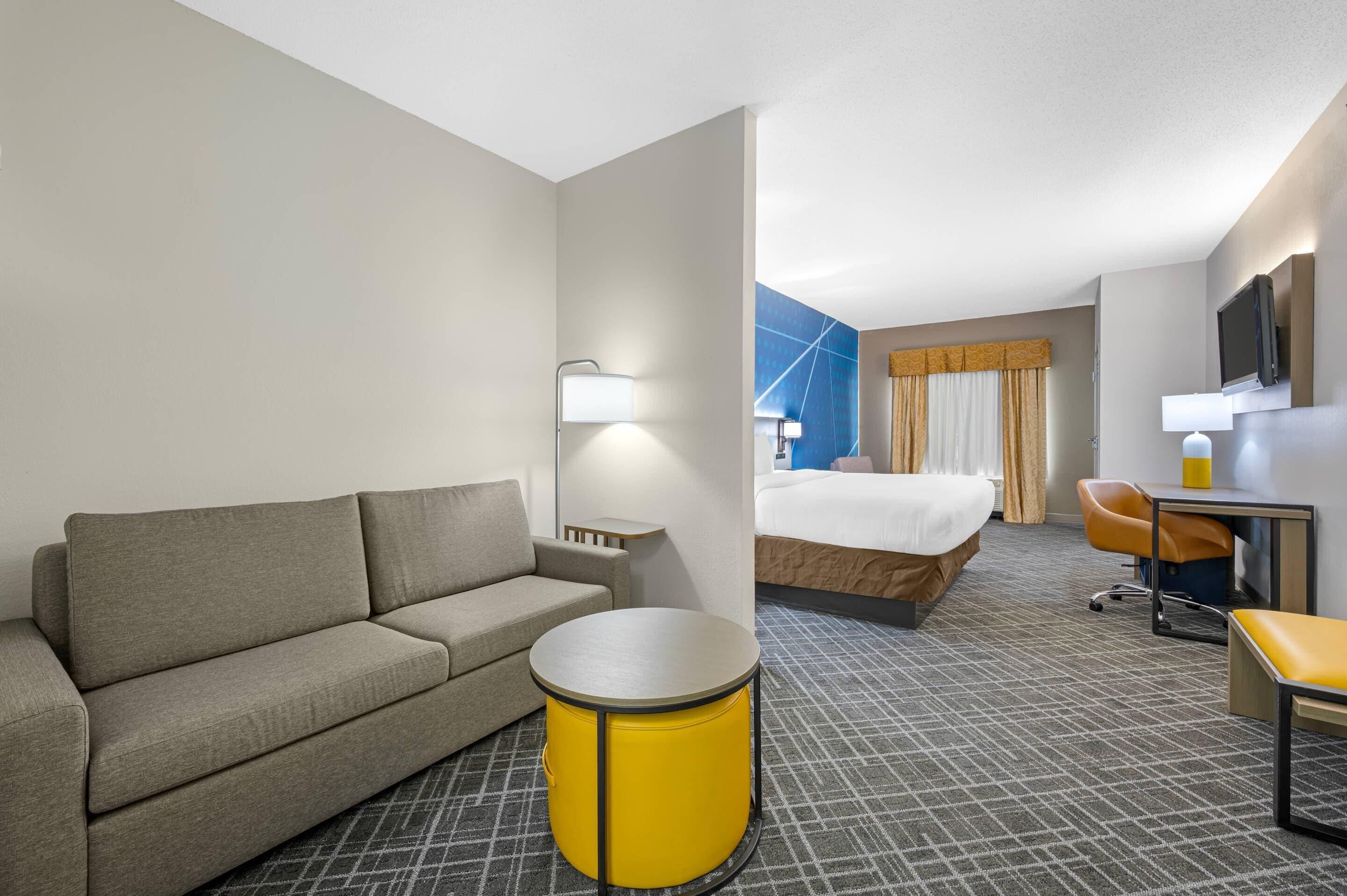 Una habitación de hotel con sofá, una mesa redonda amarilla, cama, escritorio y televisor.