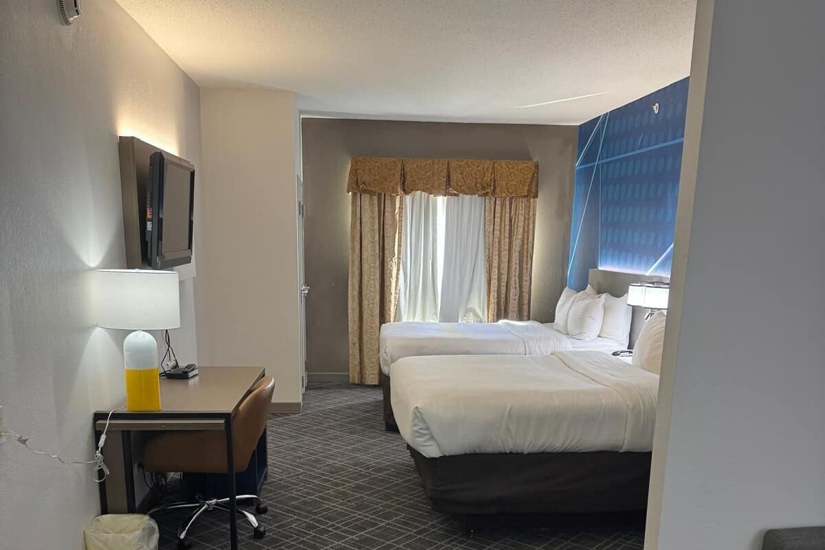 Ein Hotelzimmer mit zwei Betten, einem Schreibtisch, einem Stuhl, einem kleinen Tisch, einem Mülleimer und einem Fernseher an der Wand.