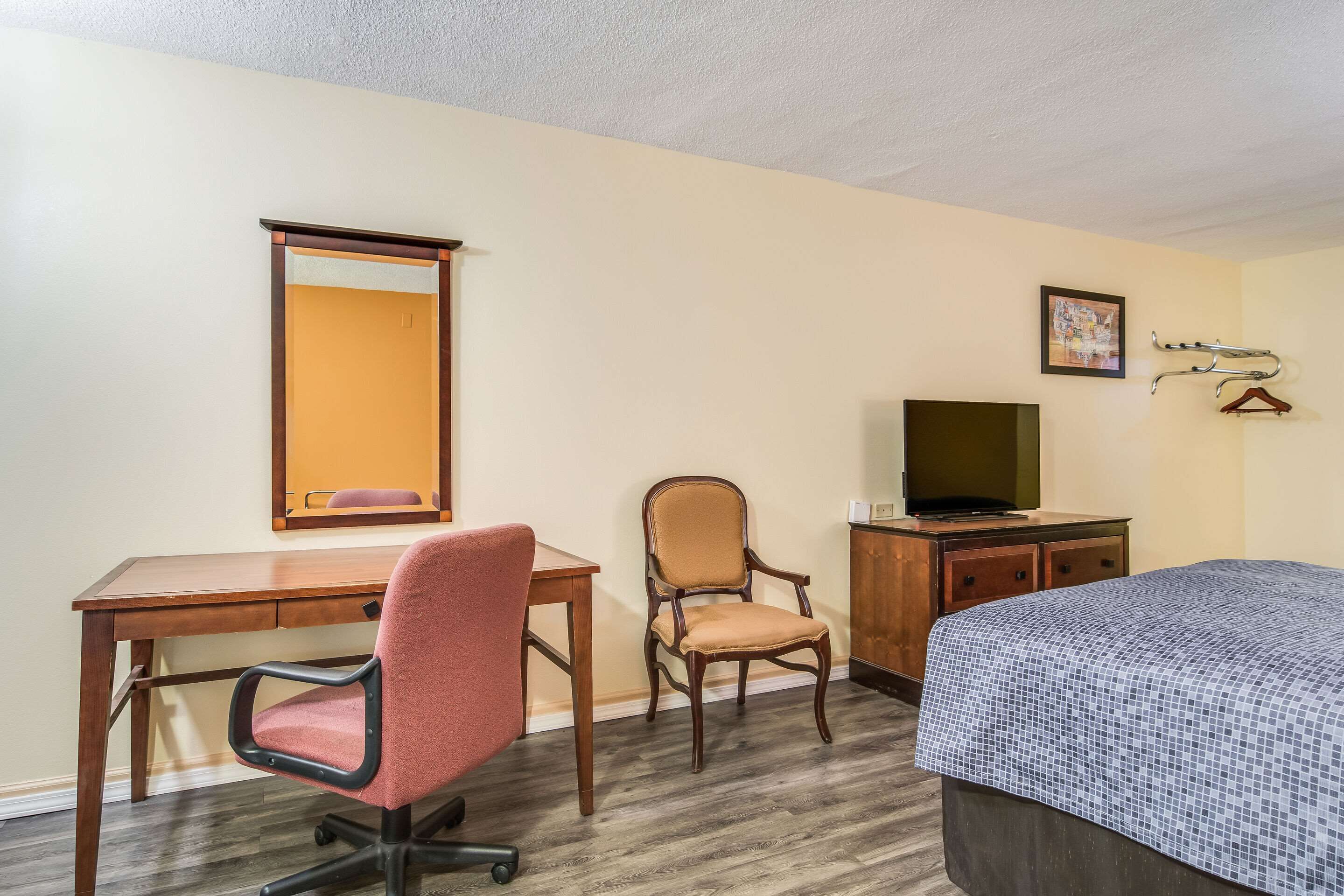Una habitación de hotel con un escritorio de madera, una silla de oficina roja, un sillón de madera, un televisor sobre un mueble de madera y una cama con colcha azul estampada.