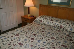 Una cama bien hecha con cubrecama floral, una lámpara de noche y un reloj digital sobre una mesita de madera.