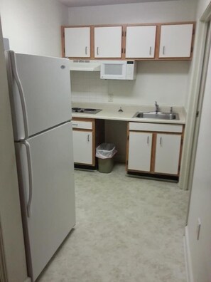 Una cocina compacta con armarios blancos, un refrigerador, un microondas, un lavamanos y un cubo de basura.