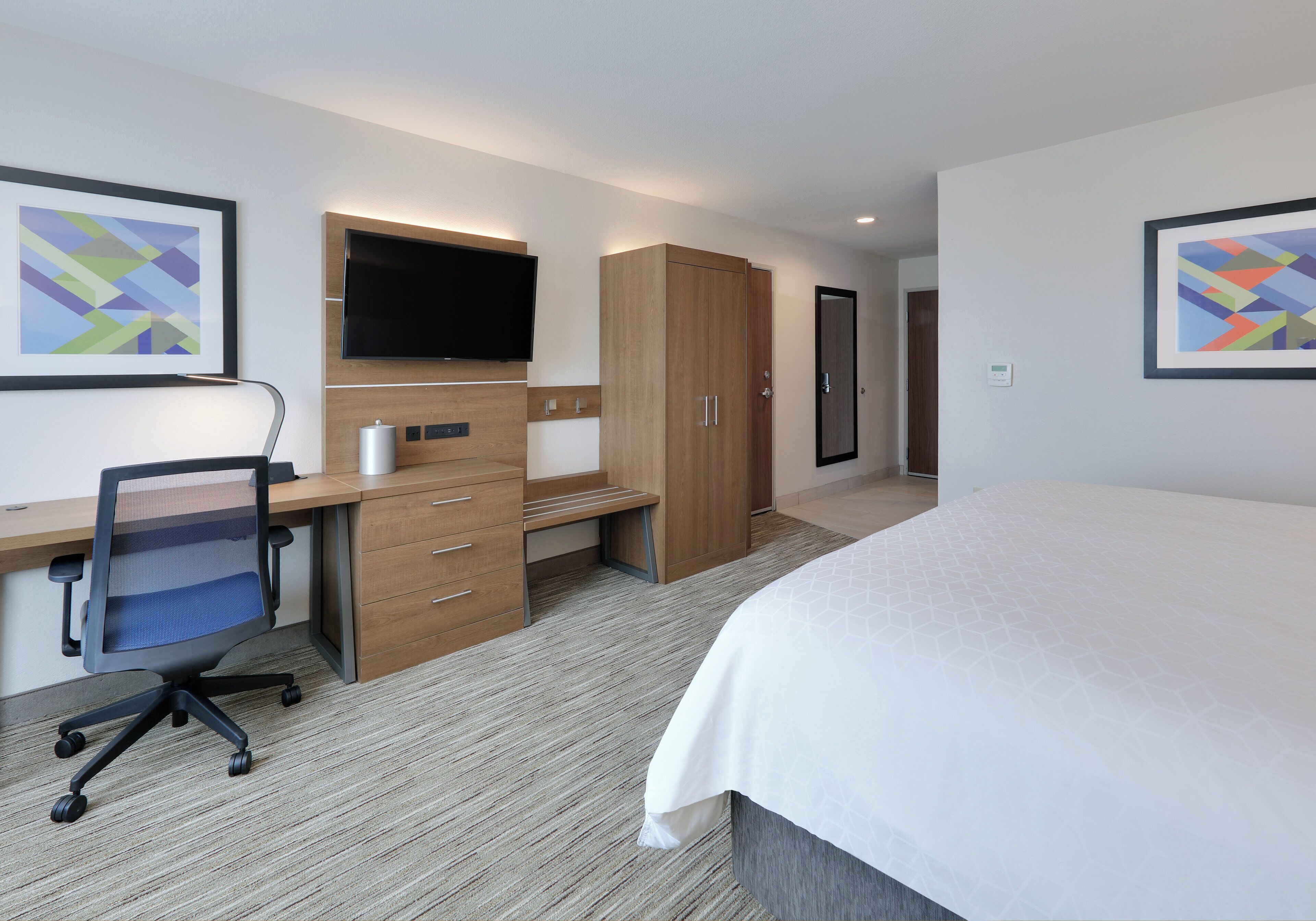 A hotel room with a bed, desk, chair, television, wardrobe, and artwork on the walls.