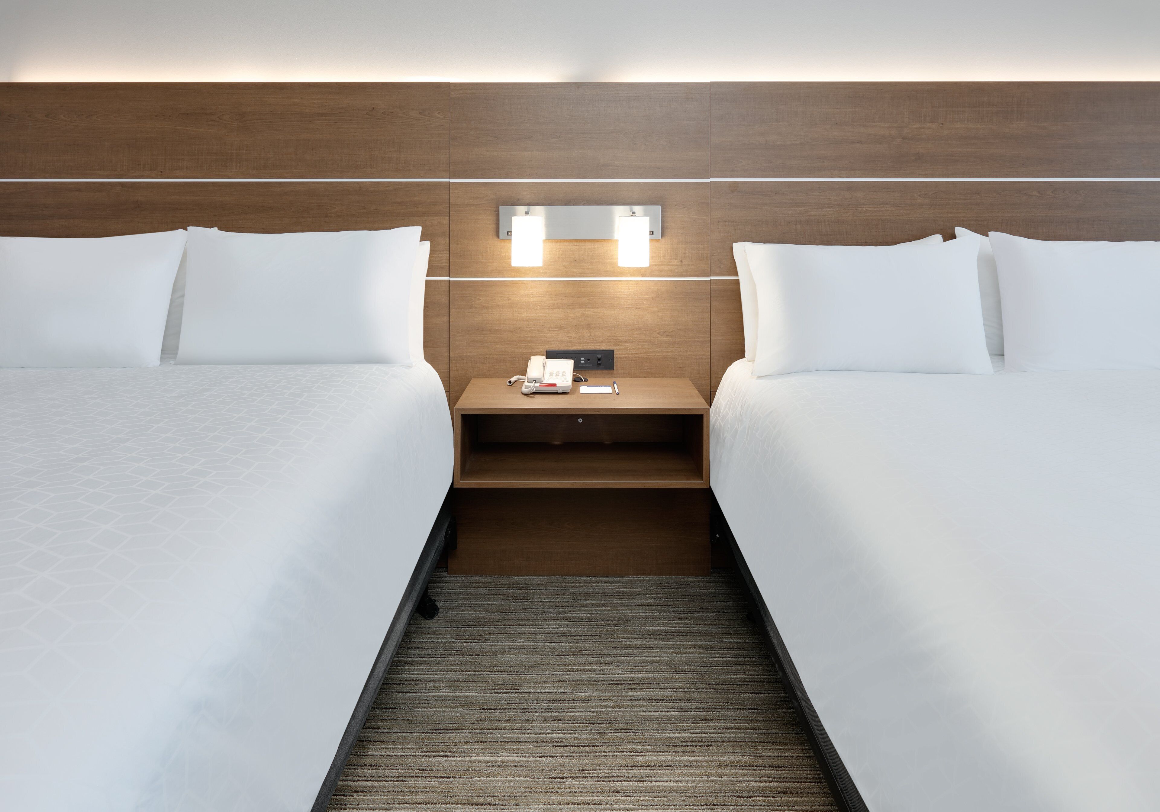 Een hotelkamer met twee bedden, een nachtkastje met een telefoon en afstandsbediening, en een houten hoofdeinde met ingebouwde verlichting.