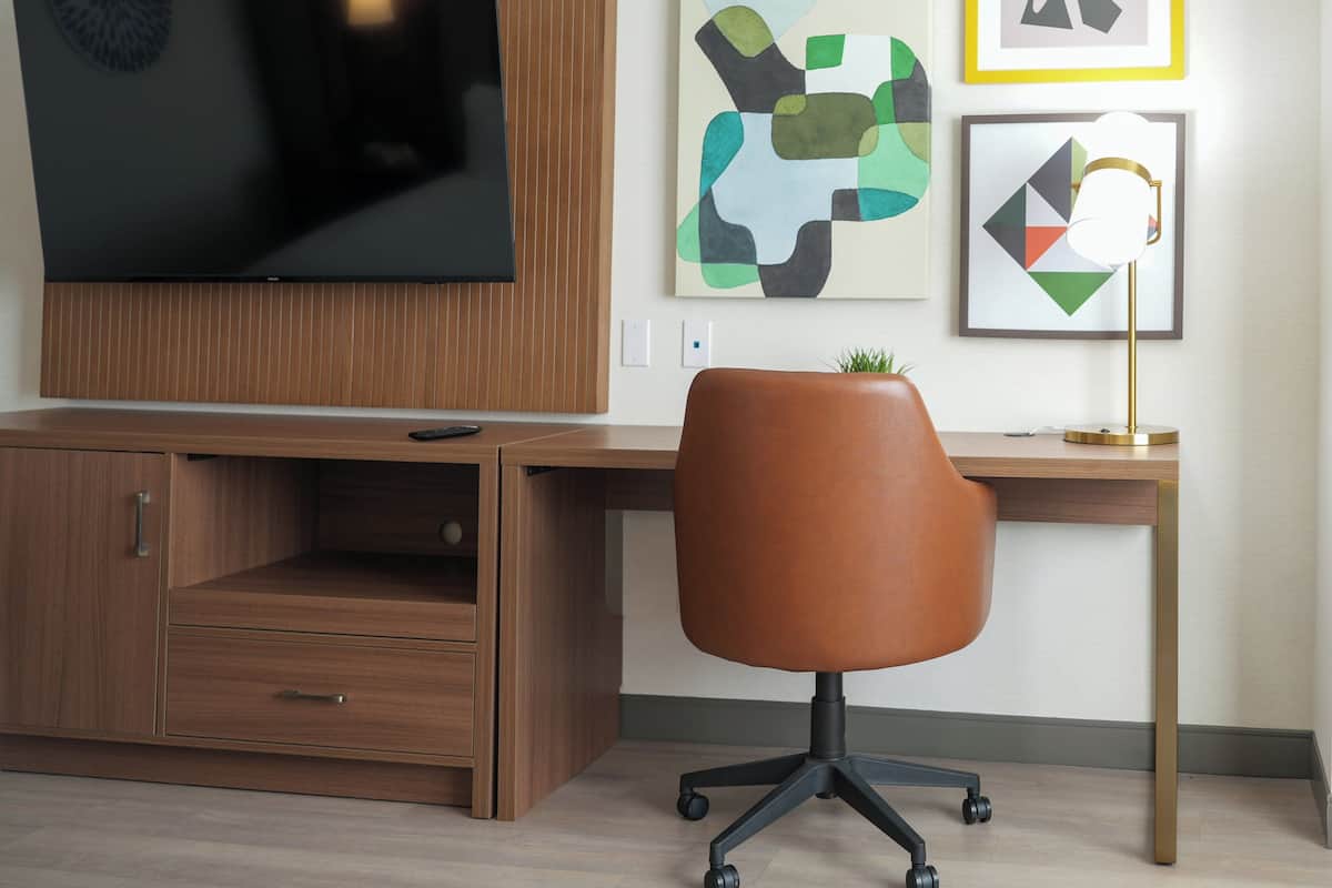 A modern office space with a wooden desk, a swivel chair, a flat-screen TV mounted on the wall, and abstract artwork on the wall.