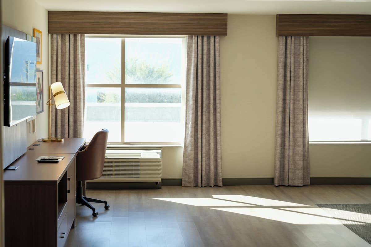 A hotel room with a desk, chair, and a large window with curtains.